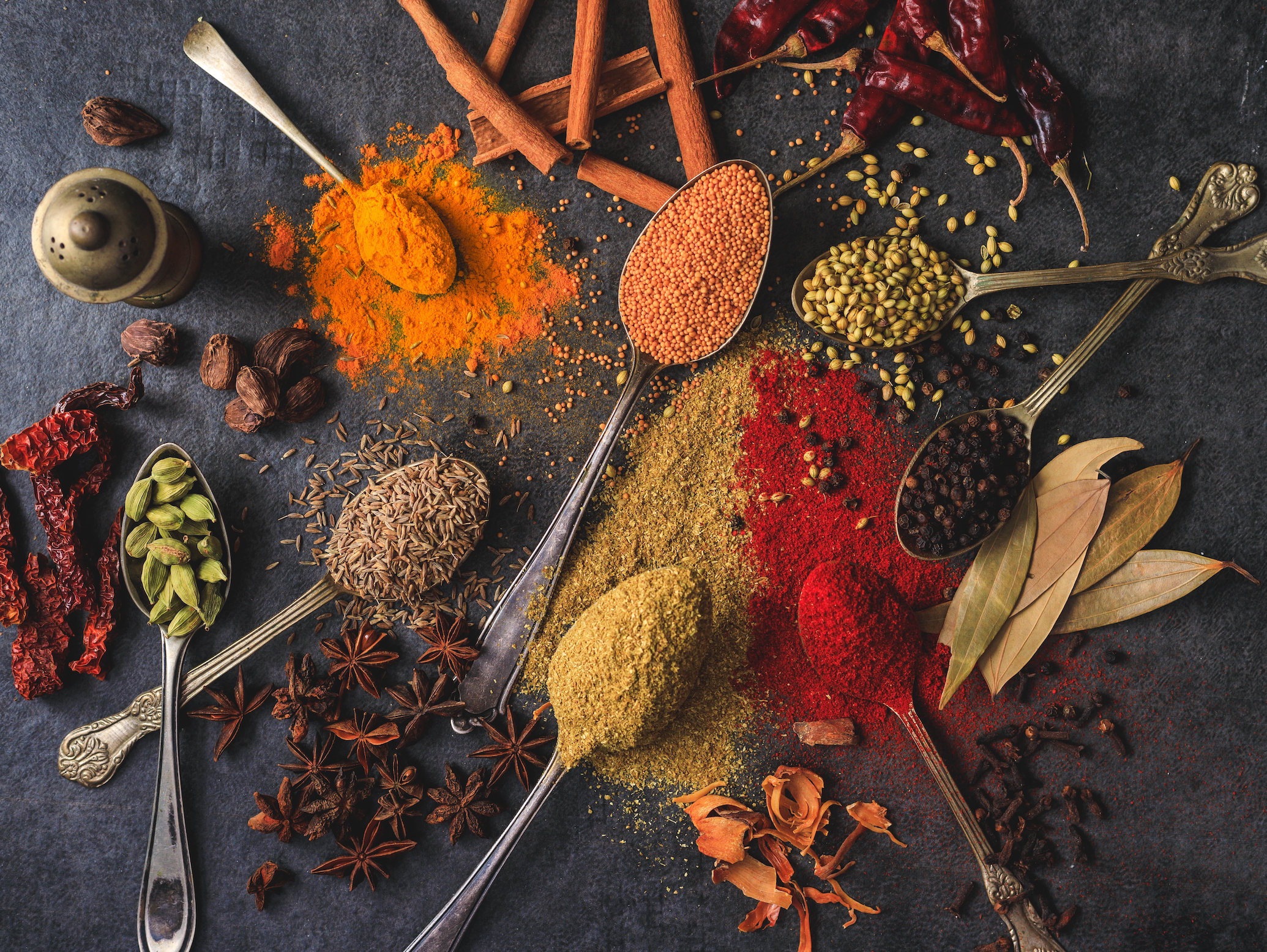 Spoonfuls of different colorful herbs and spices are on a dark wood table as examples of superfoods