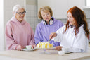 Two elderly women watching their friend slice a cake—one of the foods to avoid before plastic surgery